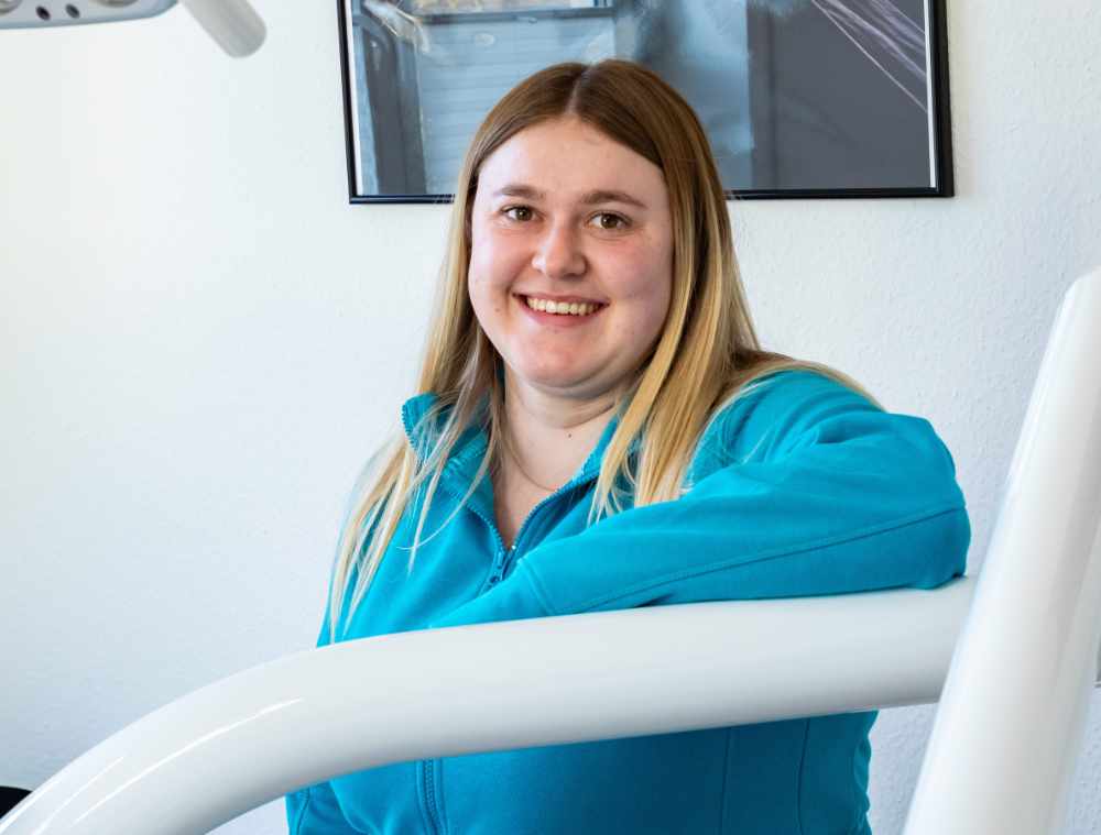 Junge Mitarbeiterin mit langen blonden Haaren und türkisfarbener Kleidung hinter einem Behandlungsgerät.